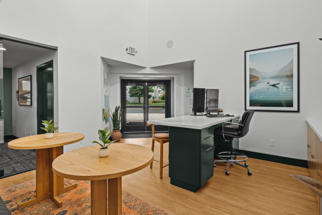 Modern leasing office lobby with wood flooring, two round tables with potted plants, a central desk with computers, a serene landscape artwork, and glass doors.