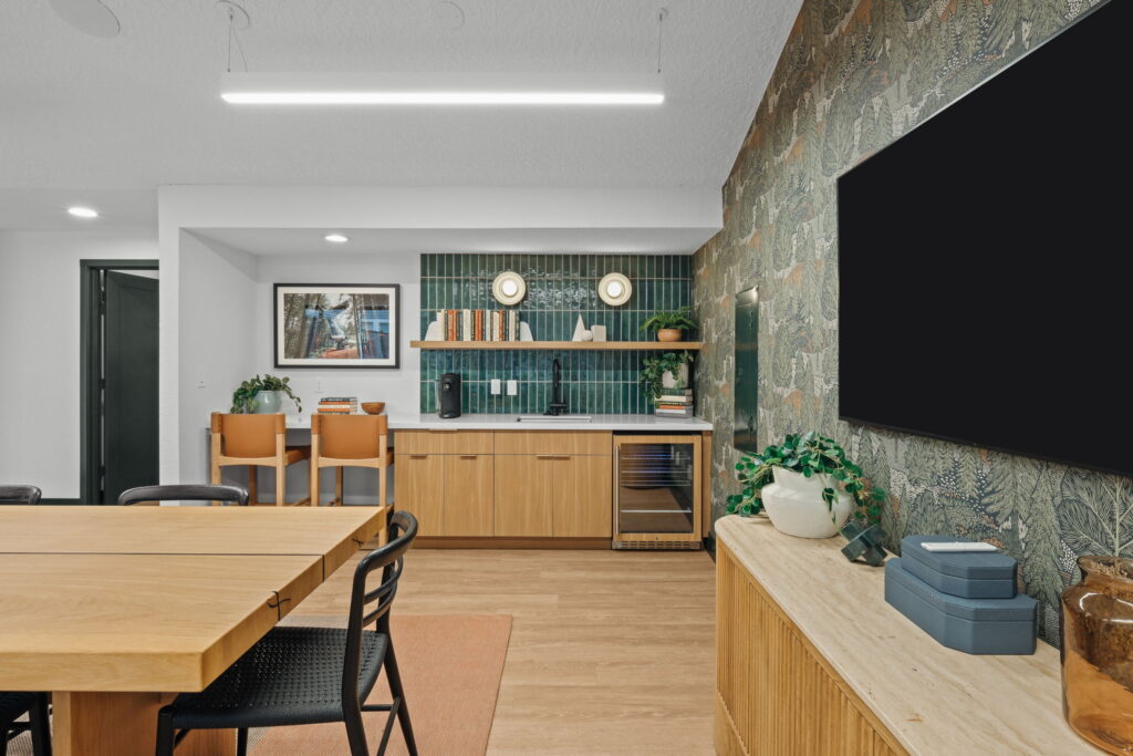 Modern leasing office dining area with kitchenette, a wooden table, wicker chairs, a green-tiled bar with books and plants, and a beige floral-patterned wall.