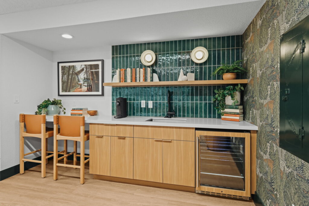 Compact kitchenette with wooden cabinets, green tile backsplash, and a shelf with books. Two bar stools face a small counter; a mini fridge is built-in.