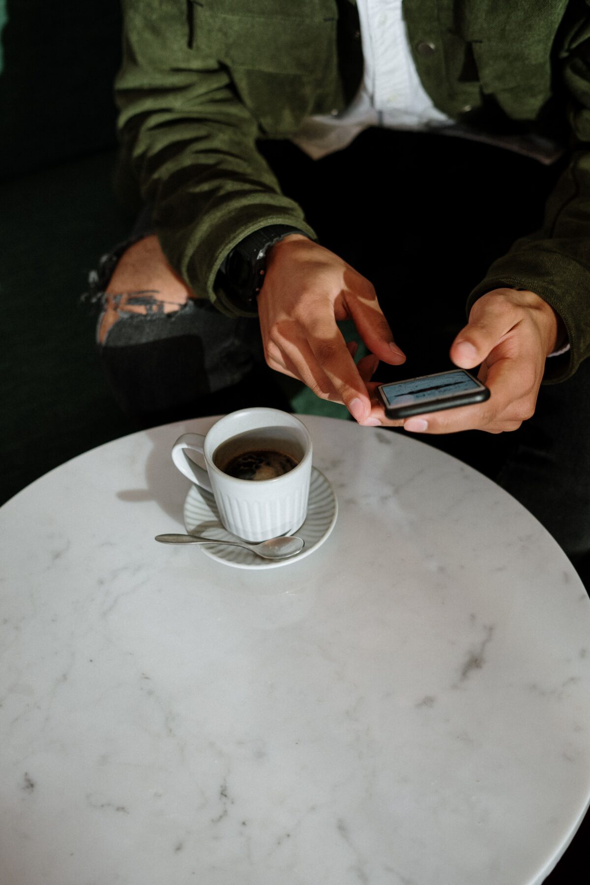 A person wearing a green jacket and ripped jeans is sitting at a round white marble table, using a smartphone. There is a cup of coffee with a saucer and spoon on the table.